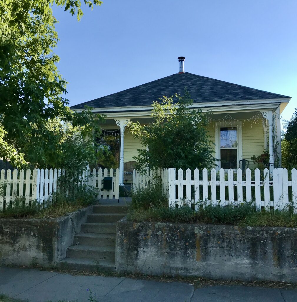 Single-story yellow house with bushes and a white picket fence. 